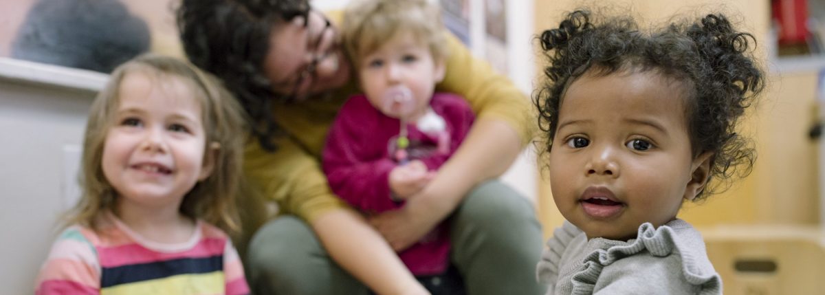 In the foreground, a toddler with curly hair looks into the camera. The background depicts an early childhood setting with an adult wearing glasses holding a baby and another toddler smiling up and outside of the camera.